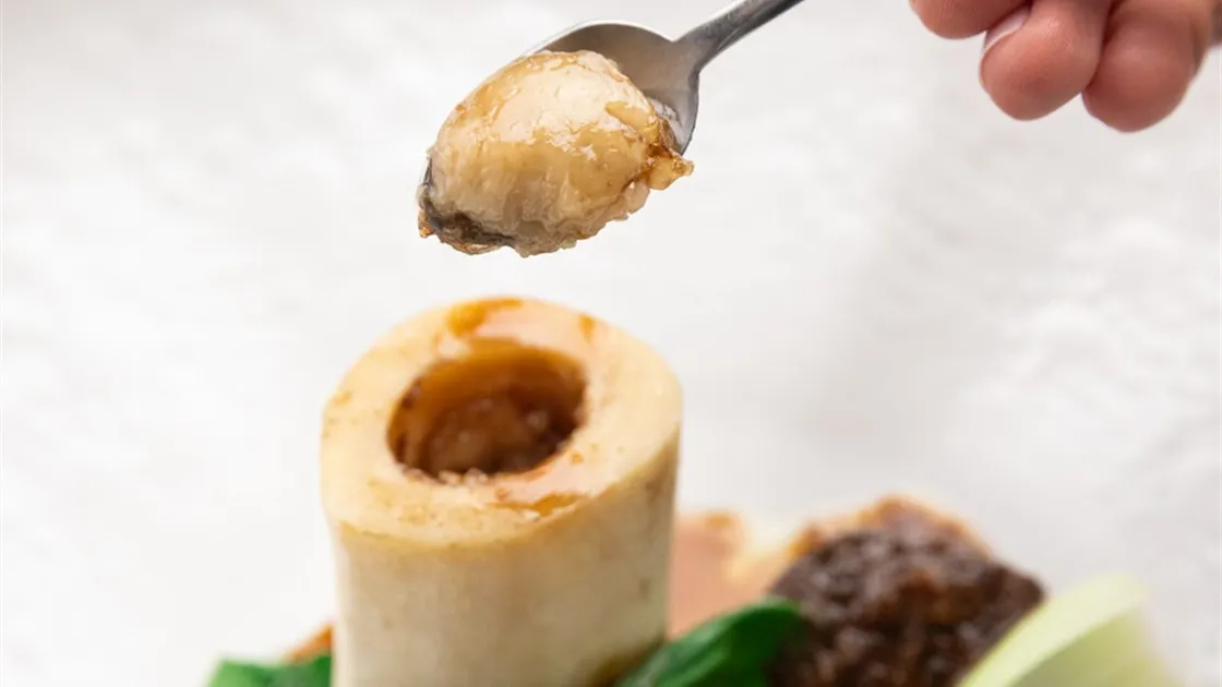 Colher retirando o tutano de um osso de ossobuco do Picchi, com pedaços de carne e folhas verdes no prato