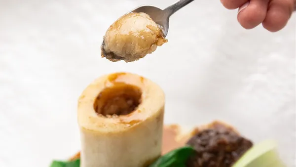 Colher retirando o tutano de um osso de ossobuco do Picchi, com pedaços de carne e folhas verdes no prato